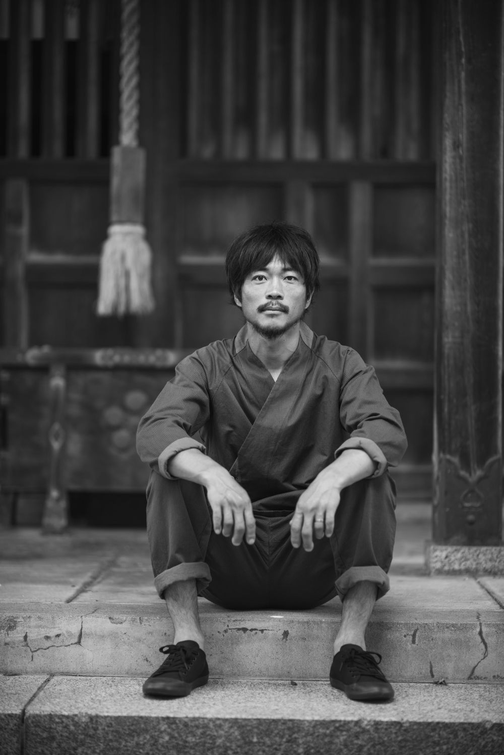 Black and white photo of someone in traditional Asian clothing sitting on stone steps by a temple entrance.