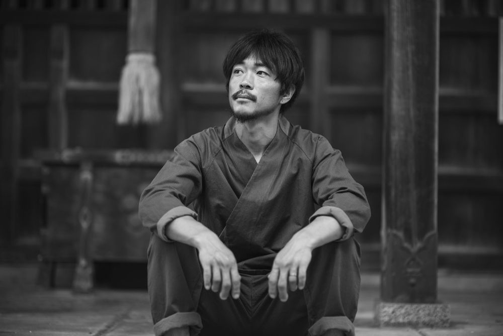 A person in traditional Japanese clothing sits on wooden steps in a contemplative black and white portrait.