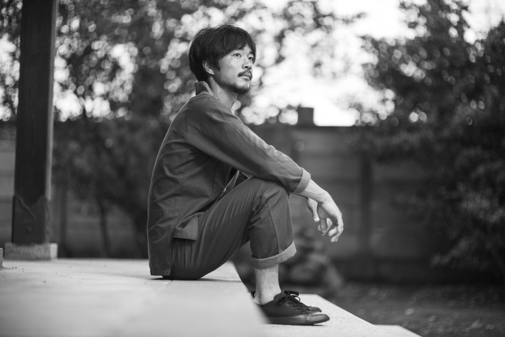 Black and white photo of someone dressed casually sitting outdoors and gazing thoughtfully into the distance.