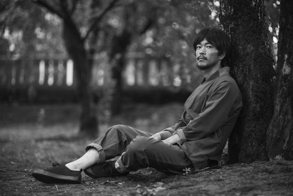 A person in dark casual attire sits against a tree trunk on the ground in a tranquil black and white setting.