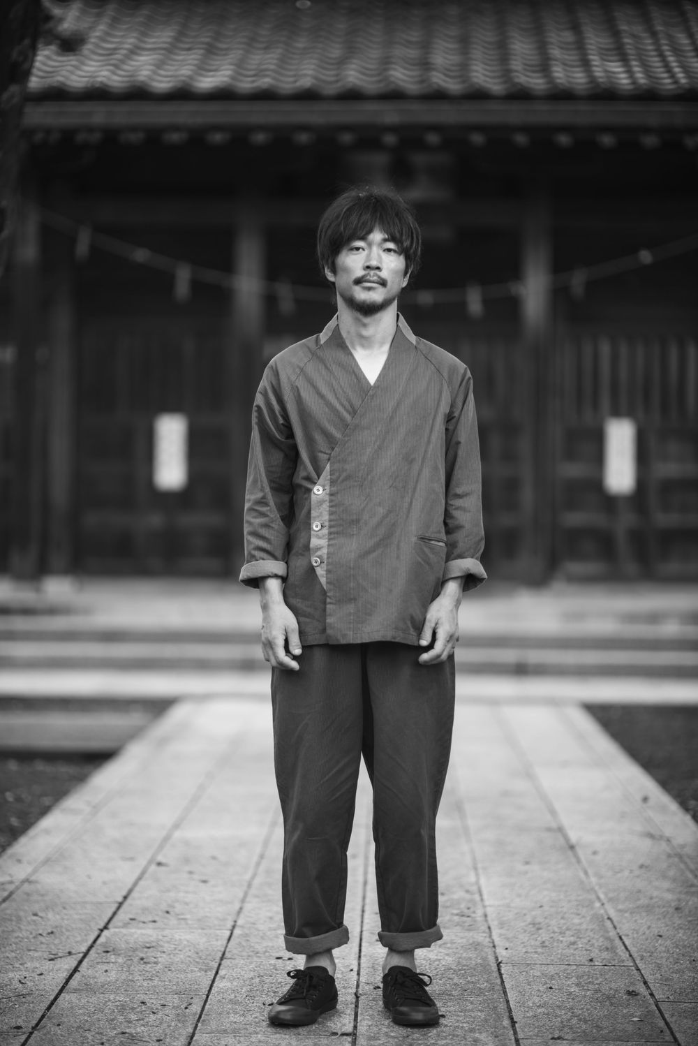Black and white portrait of someone wearing traditional Japanese clothing standing in front of a temple building.