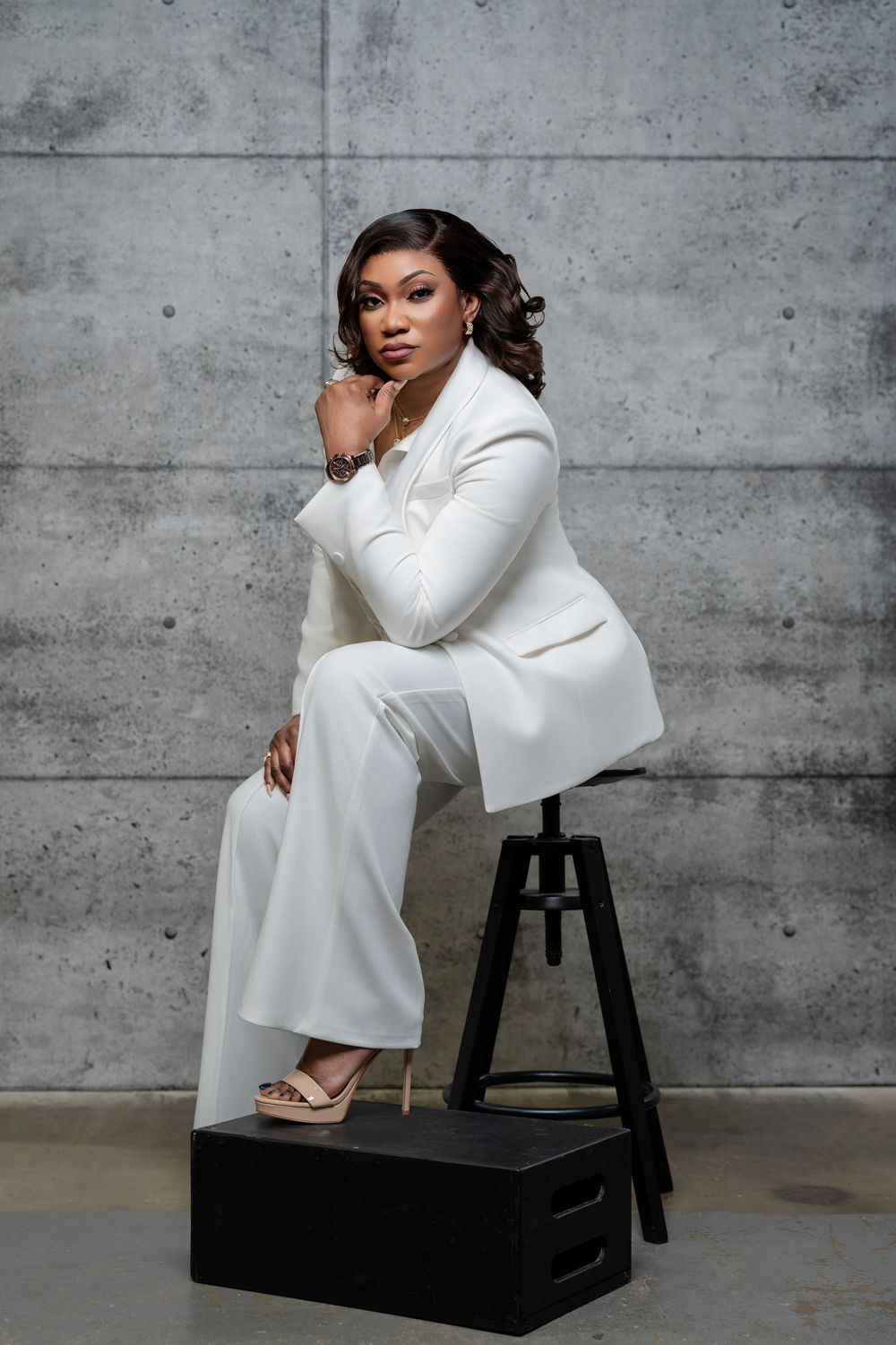 Professional portrait of a model in a crisp white pantsuit posing on a stool against an industrial concrete backdrop.