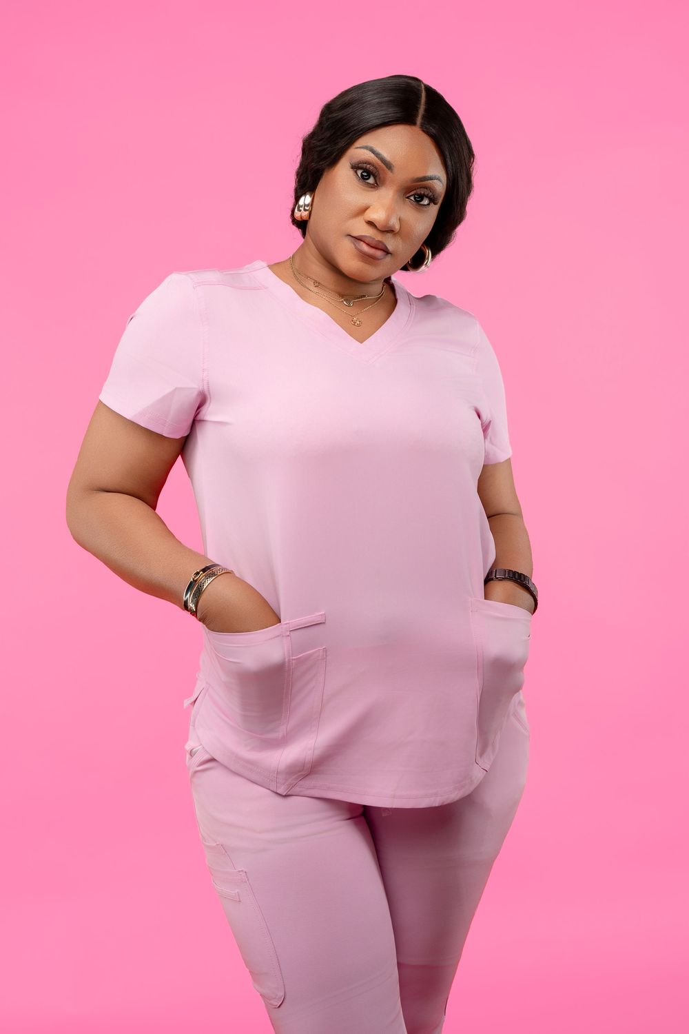 Healthcare worker in pink scrubs stands confidently against pink background with hands in pockets.