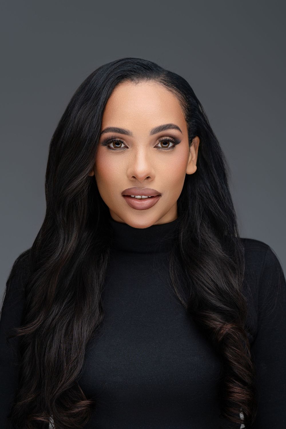 Professional portrait of someone with long dark wavy hair wearing a black turtleneck against a gray background.