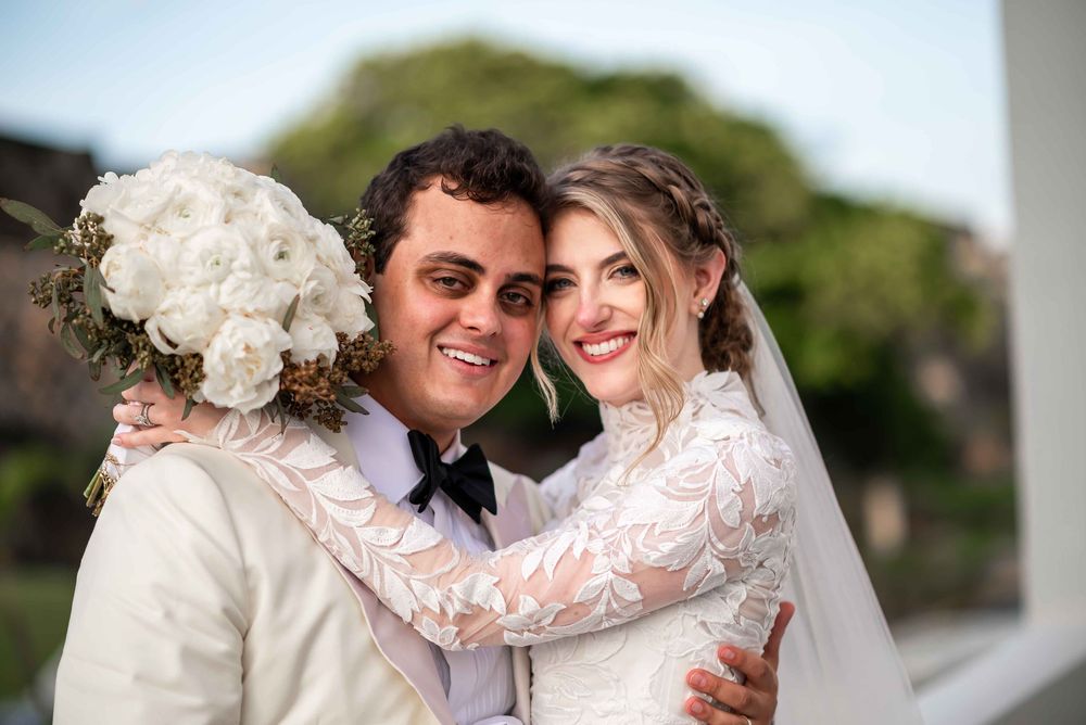 A happy couple embraces in formal wedding attire with lace details and a white floral bouquet outdoors.