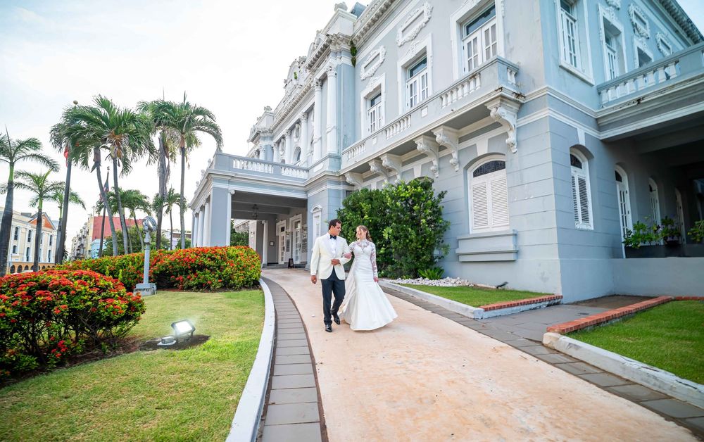Elegant couple walks along curved pathway leading to grand white colonial mansion with palm trees and landscaped gardens.