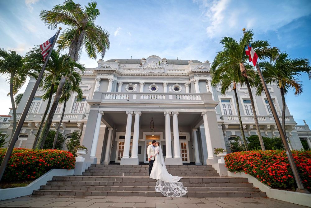 A grand white colonial mansion with tall palm trees and ornate columns creates a majestic wedding venue backdrop.