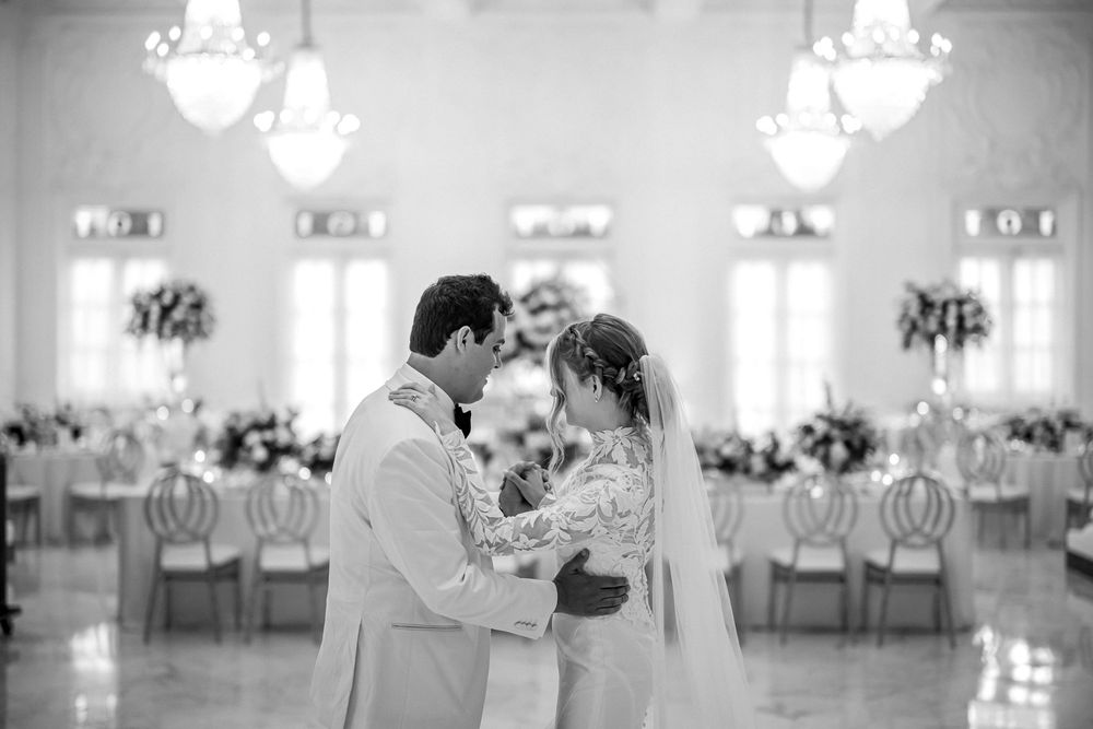 Black and white photo of a couple sharing an intimate moment in an elegant ballroom with chandeliers and floral decor.