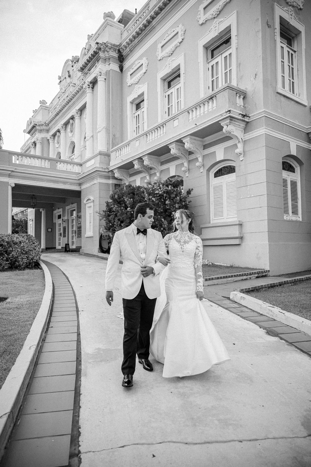 Elegant couple walk along curved path outside historic mansion building in black and white photography.
