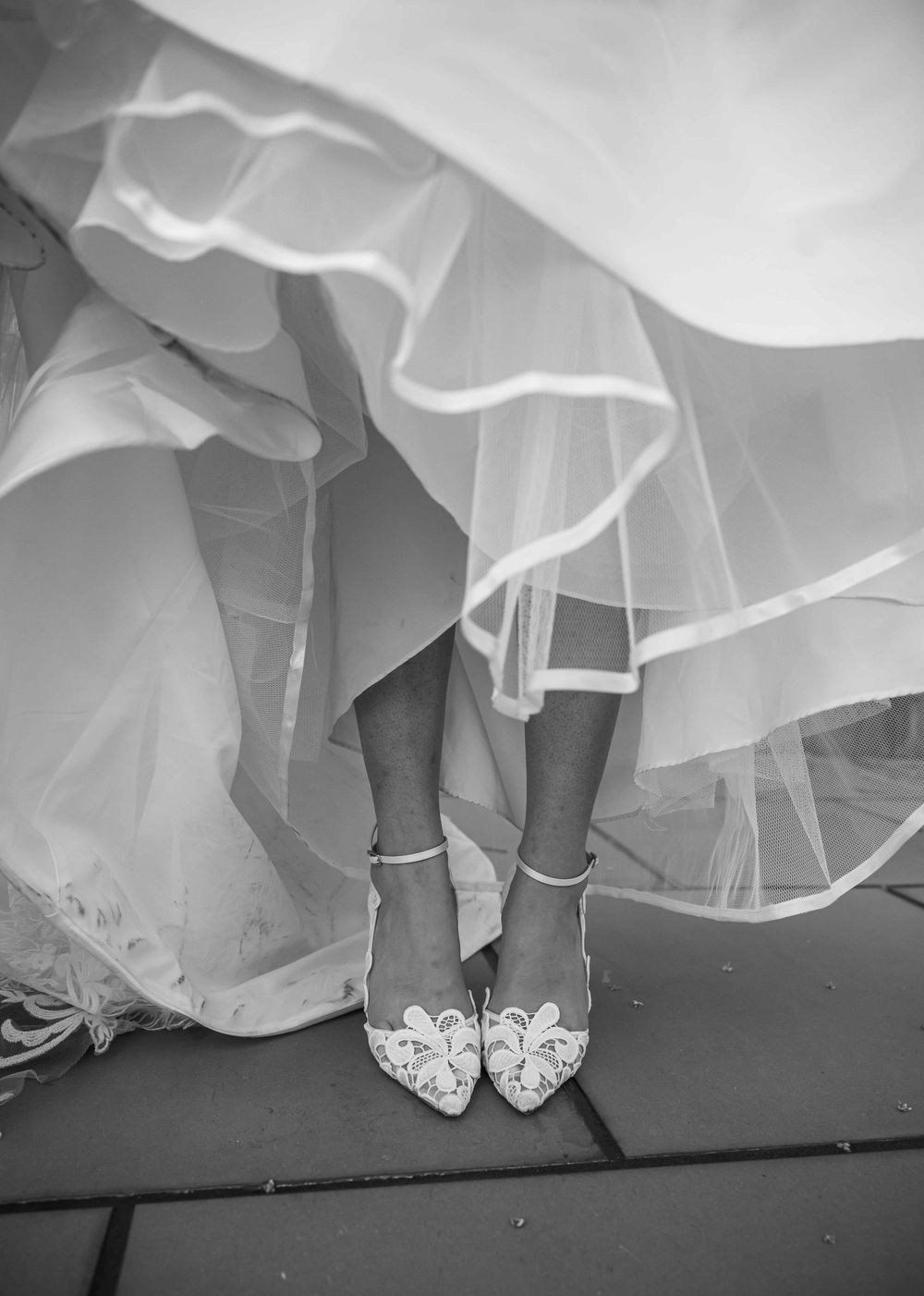 A close-up black and white photo of ornate bridal shoes with bows beneath layers of flowing wedding dress fabric.