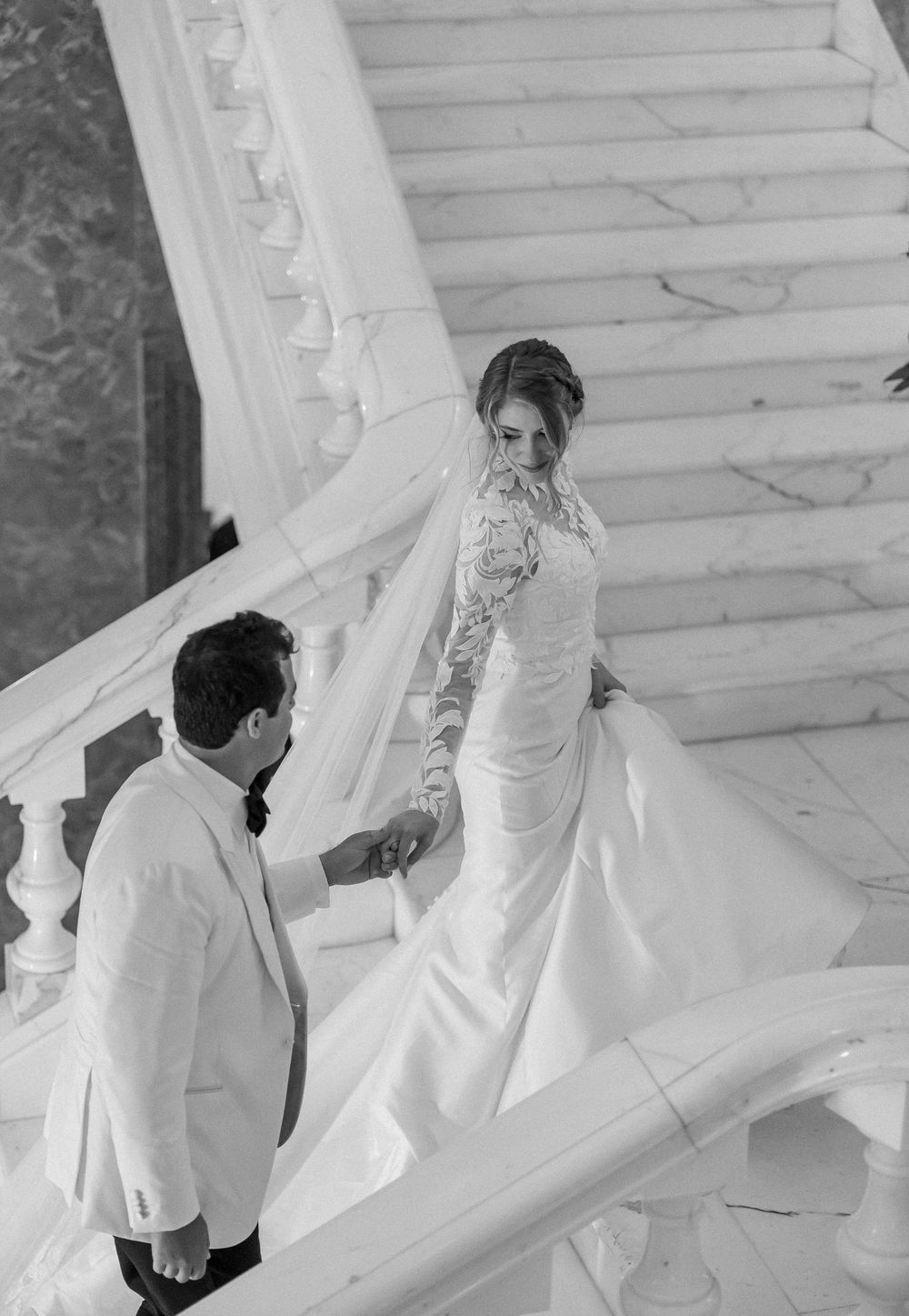 A black and white wedding photo captures a couple ascending elegant marble stairs in formal attire and a flowing veil.