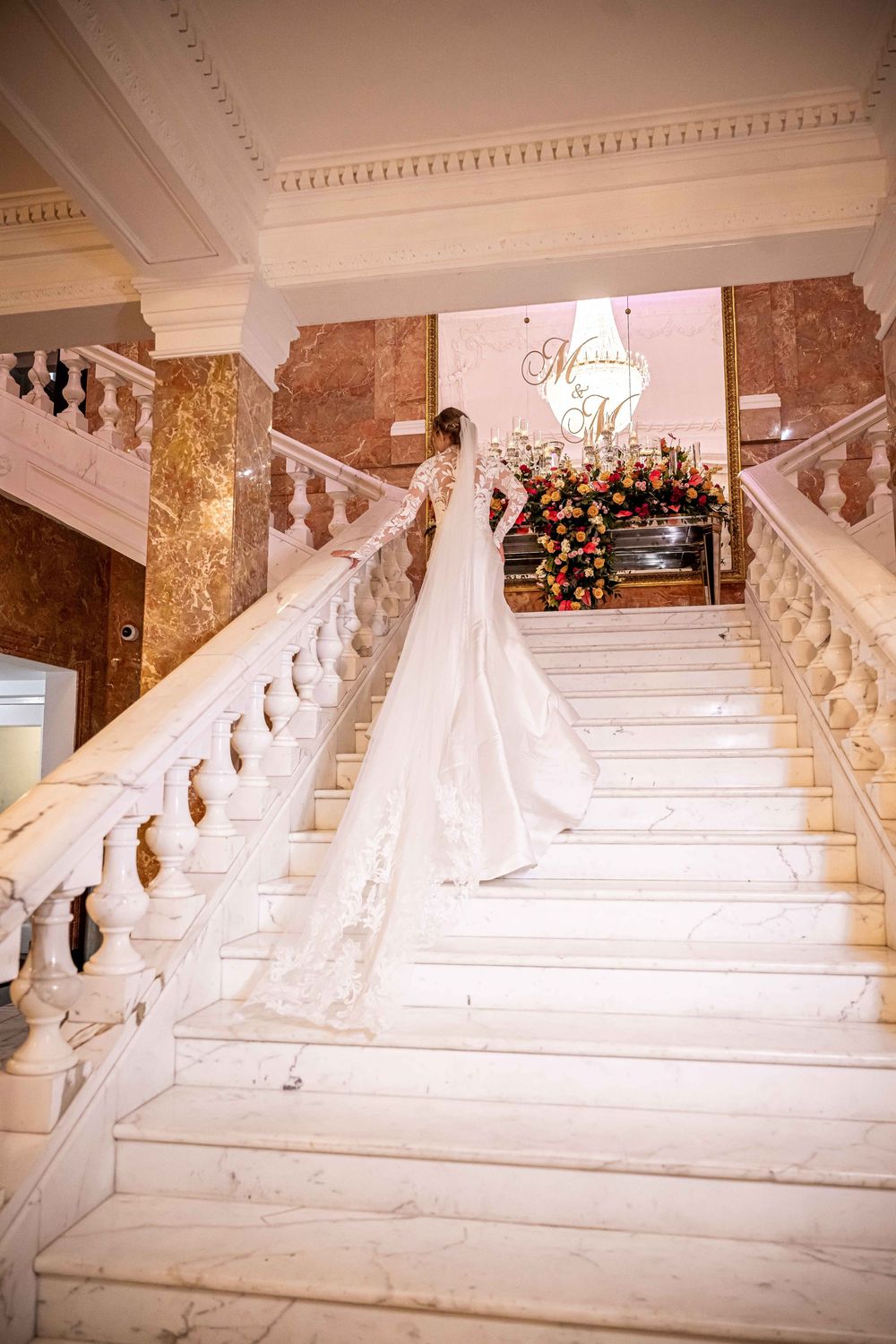 A bride in an elegant white gown with a long flowing train stands on a grand marble staircase with floral decorations.