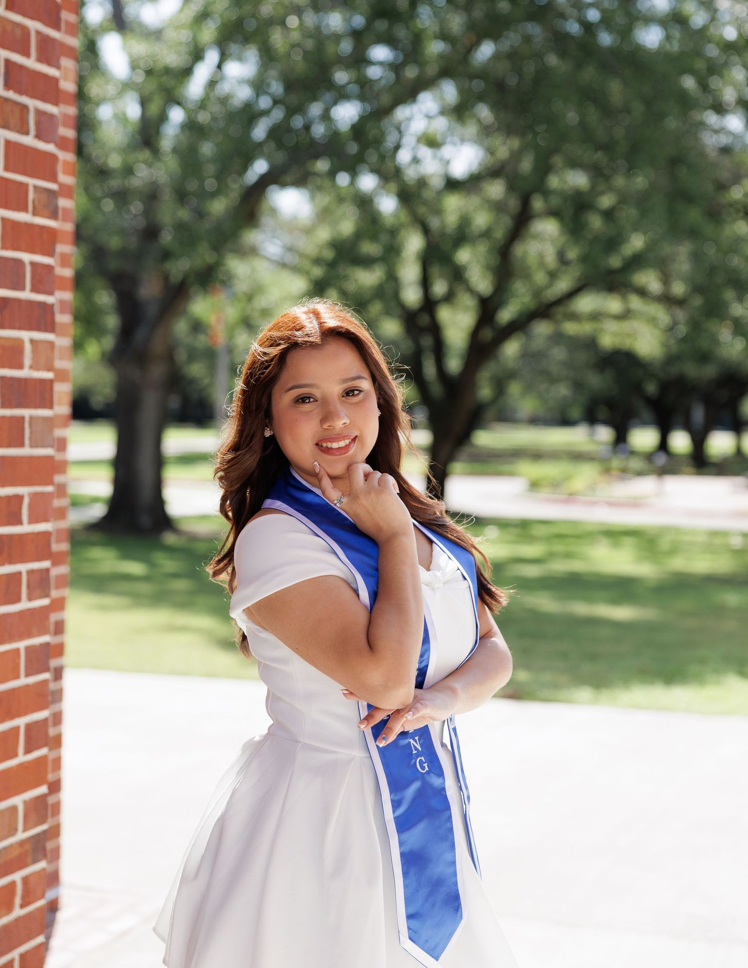 Graduate in cap and gown poses for commemorative photos against brick wall and greenery in outdoor setting.