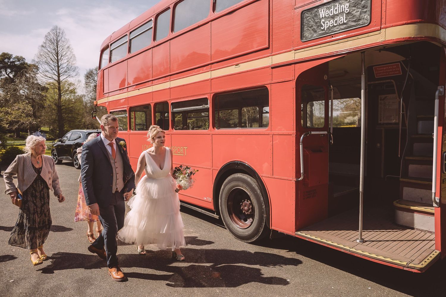 Wedding guests board a vintage red double decker bus for transportation on a sunny day.