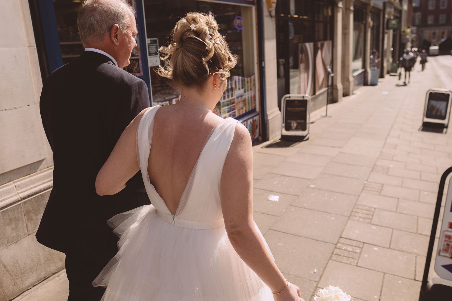 The bride and father of the bride walk towards The Botanist with the bride wearing a white dress with plunging v-back.