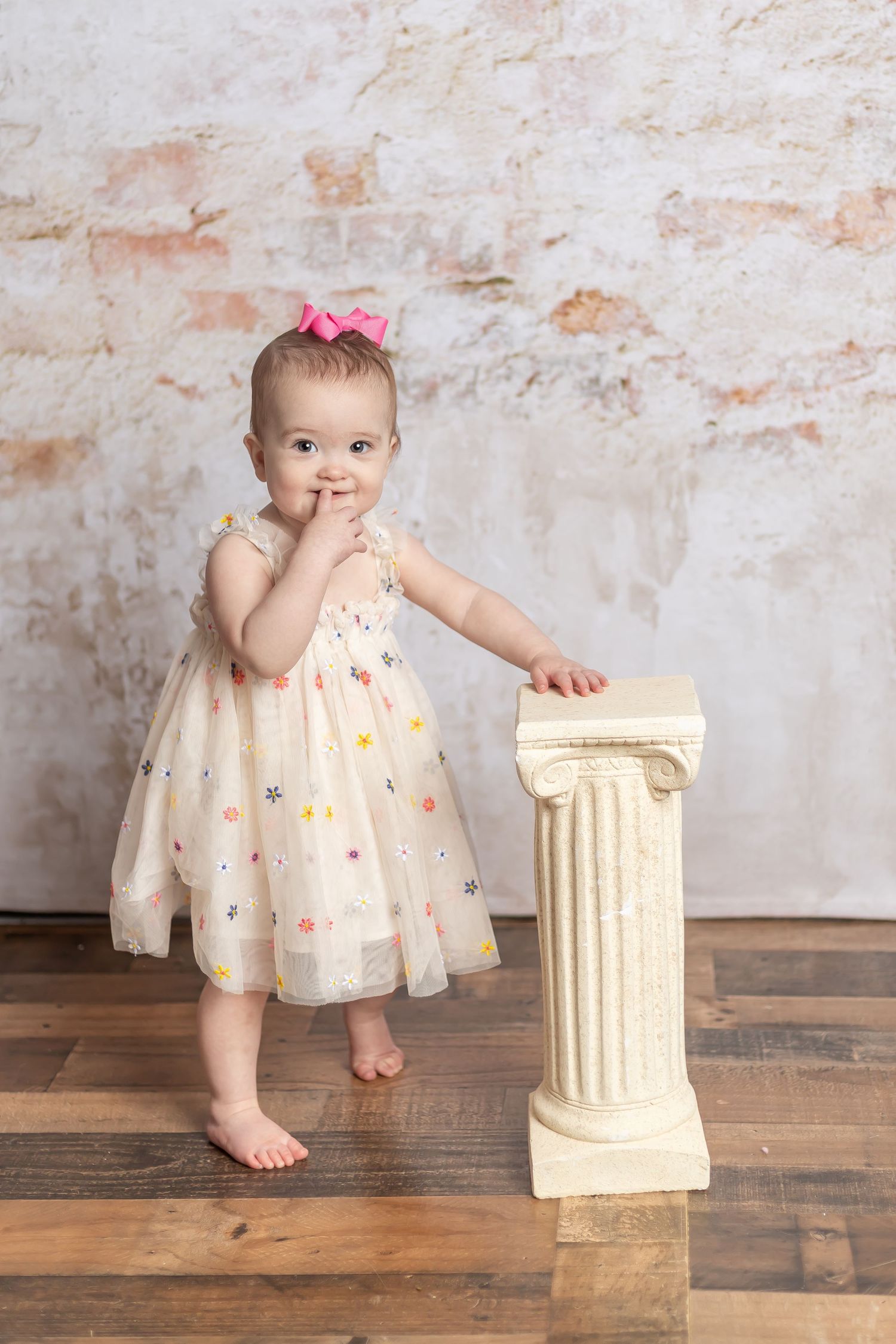 A small child in a floral dress stands next to a decorative white column against a textured wall background.