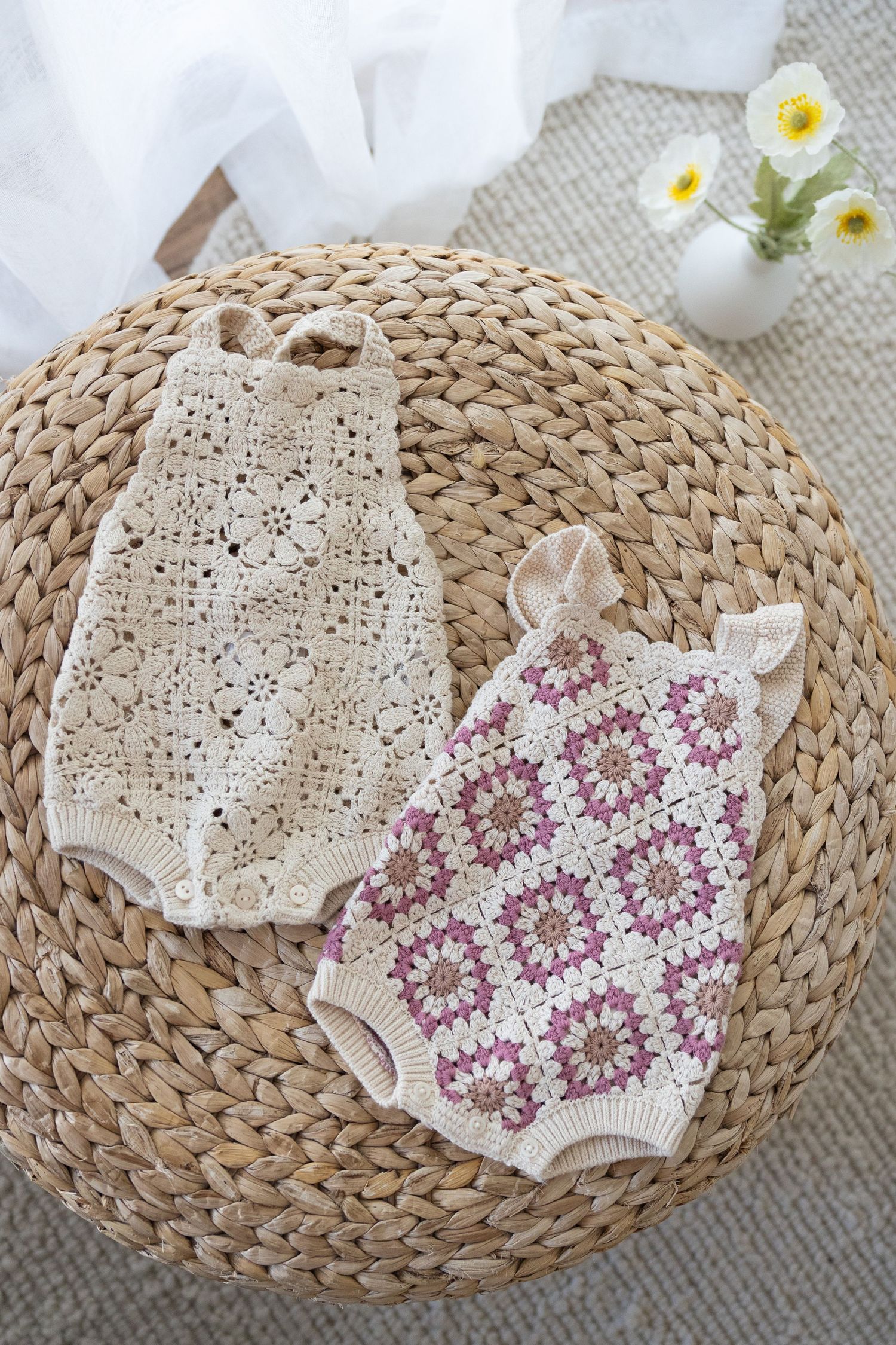 Two crocheted baby rompers in cream and pink floral patterns displayed on a round woven rattan mat with white flowers nearby.