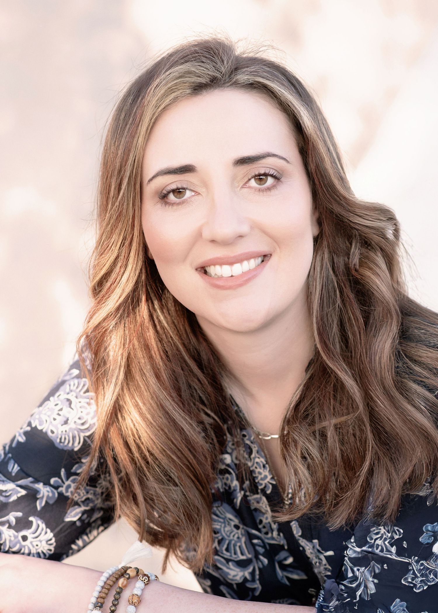 Portrait of a smiling person with long wavy brown hair wearing a dark floral print top against a soft pink background.