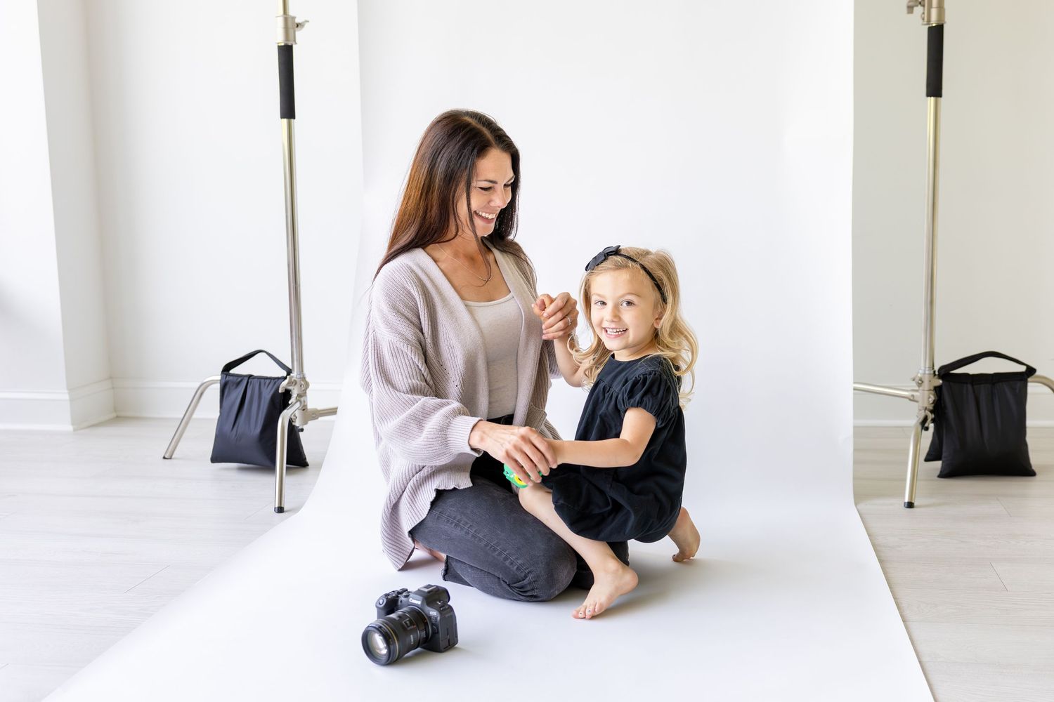 A mom and daugther sitting together and smiling on a white studio backdrop with lighting and photography equipment visible.