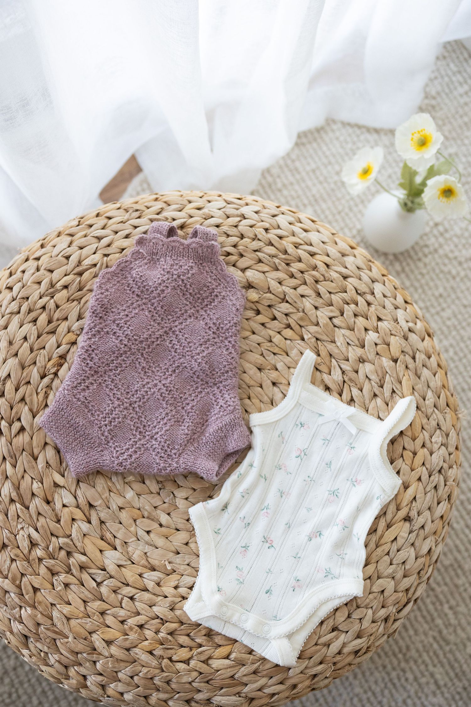 Baby bodysuits in lilac and white laid on woven rattan mat with small white flowers in vase.