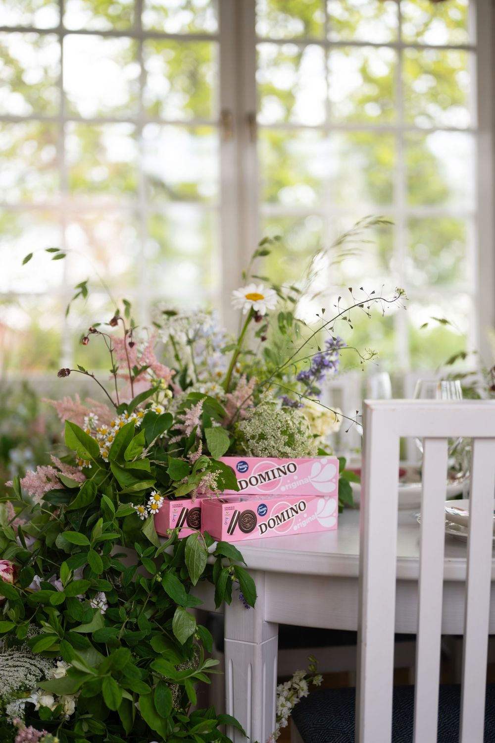 Pink sunglasses and delicate floral arrangements adorn a white wooden chair by a bright sunlit window.