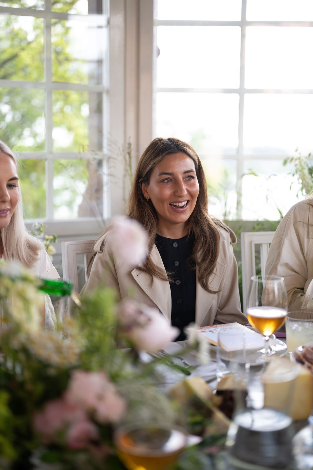 Friends enjoying drinks and conversation at a bright sunlit table decorated with flowers during a social gathering.