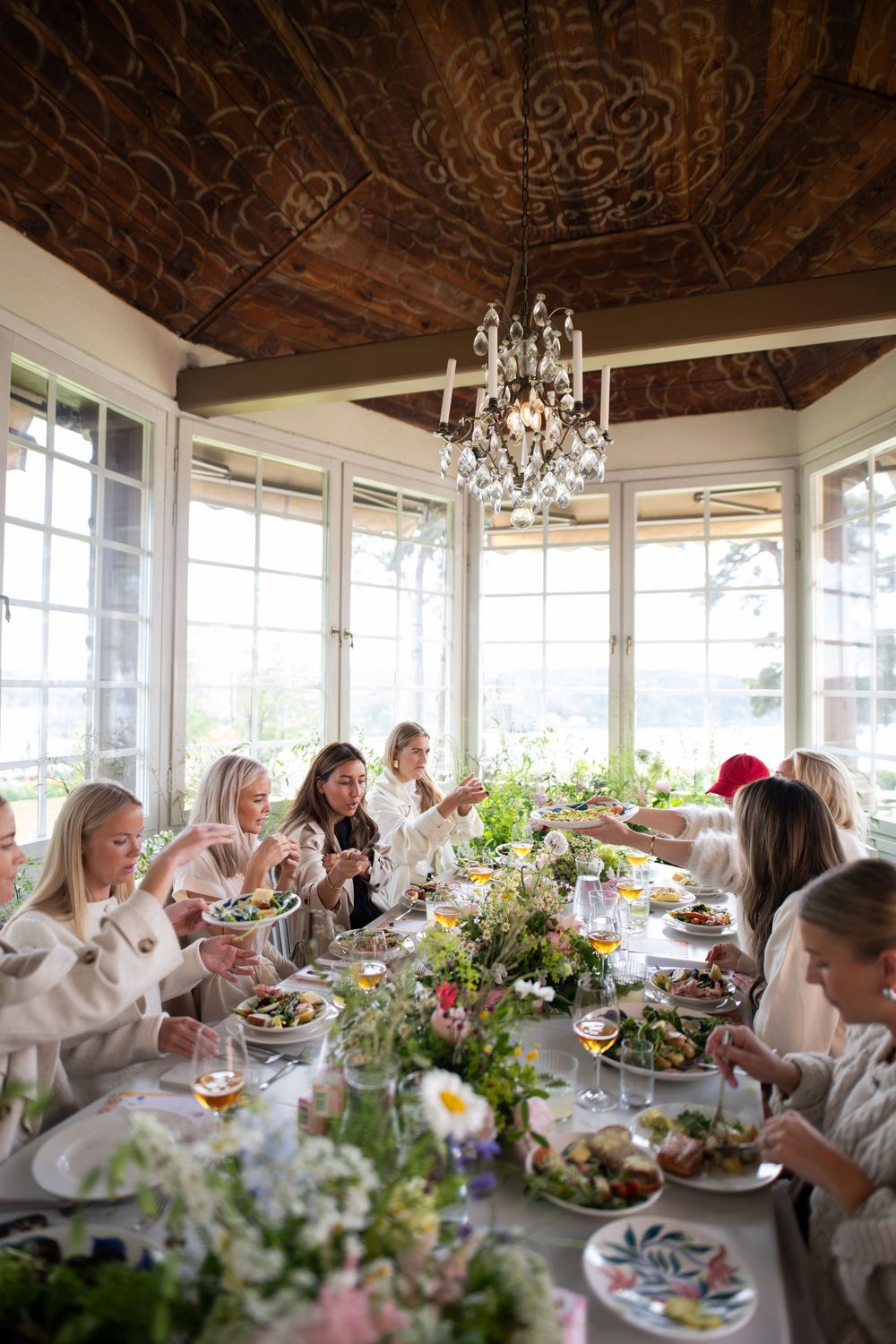 Diners enjoy an elegant lunch around a table decorated with floral arrangements under an ornate wooden ceiling with chandelier.