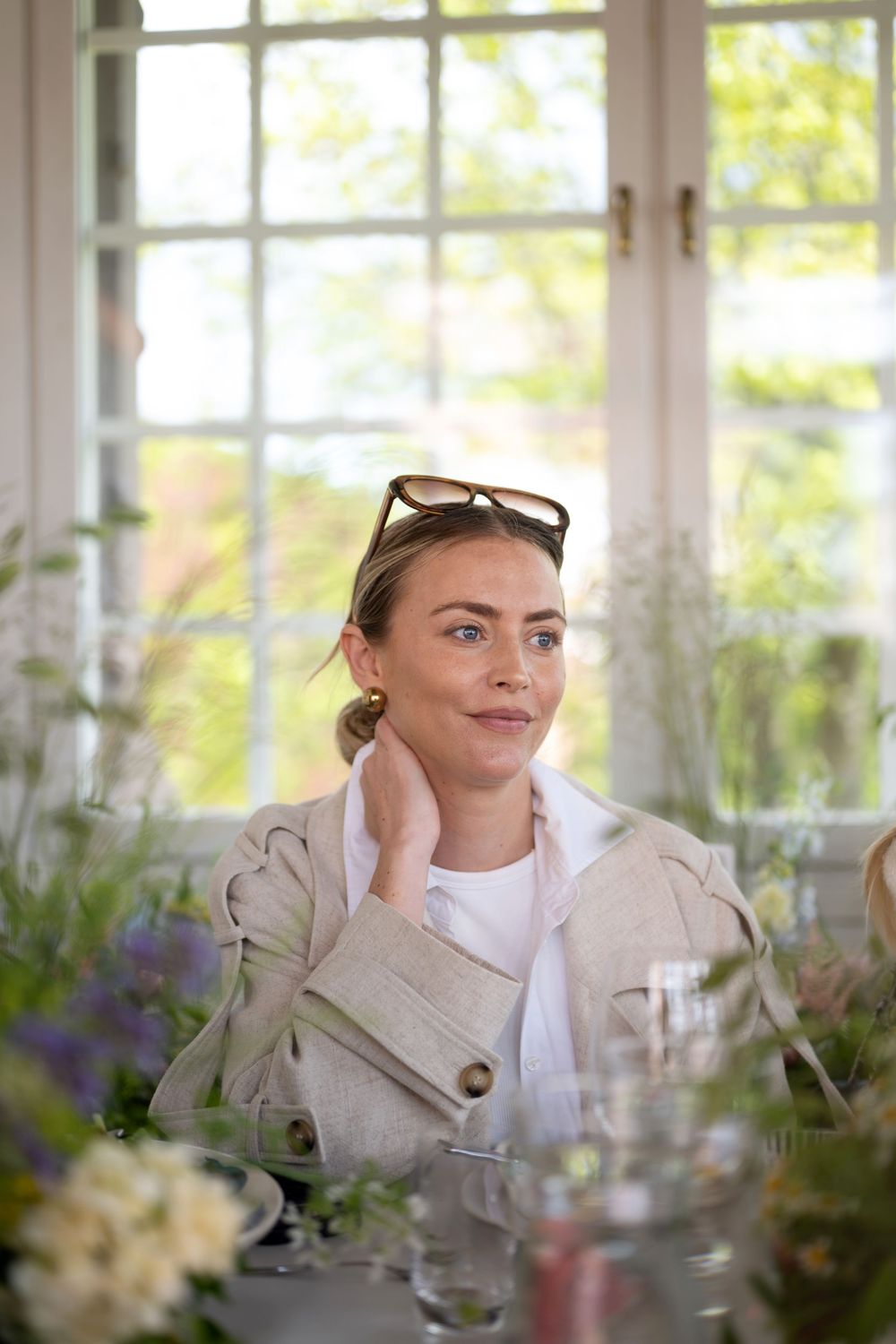 Relaxed person in beige trench coat seated at table with flowers and sunlit windows in background.