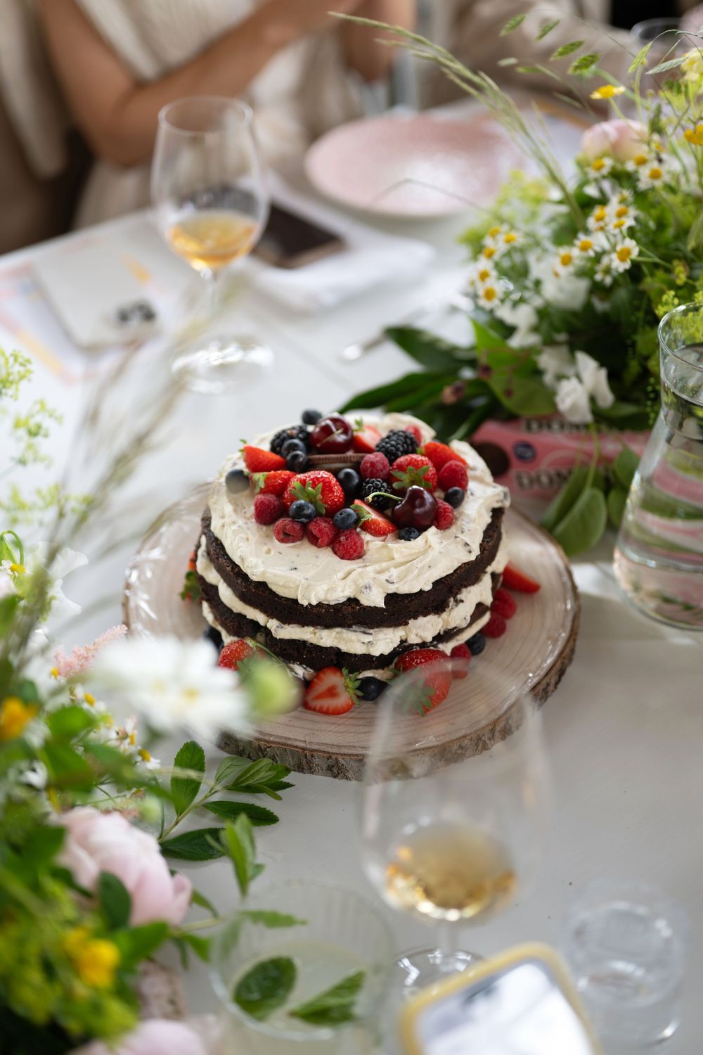Elegant chocolate layer cake topped with fresh berries and whipped cream on a decorated table with wine glasses and flowers.