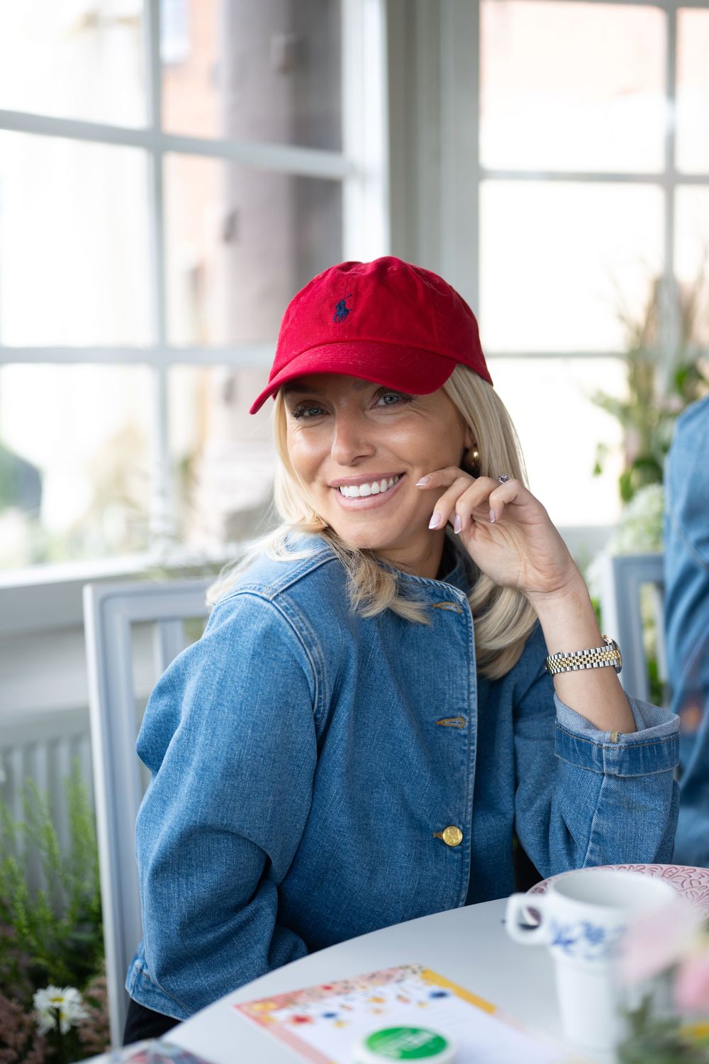 A person wearing a red baseball cap and denim shirt sits at a table with a bright smile.