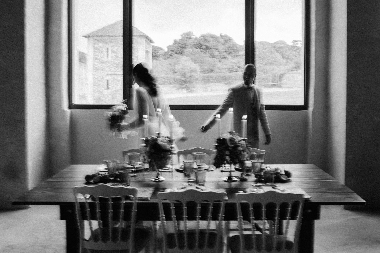 Black and white photo of bride and groom standing at a decorated wedding reception table by large windows.