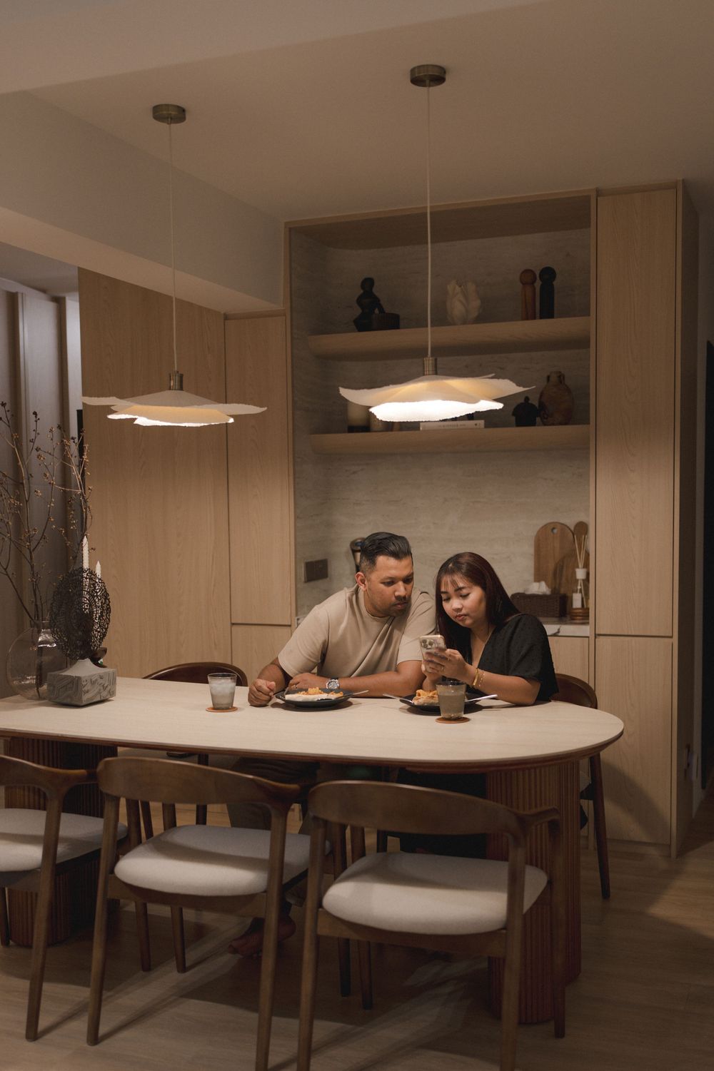 Two people sharing a cozy moment at a modern dining table with pendant lights and built-in shelving in warm lighting.