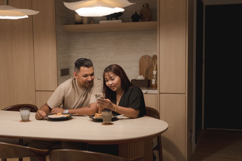 A couple smiles while looking at a mobile phone together at a dining table under pendant lights.