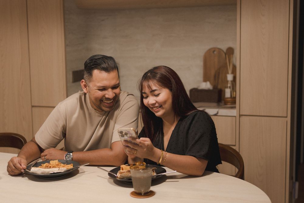 Two people share a laugh while looking at a mobile phone during a meal at home.