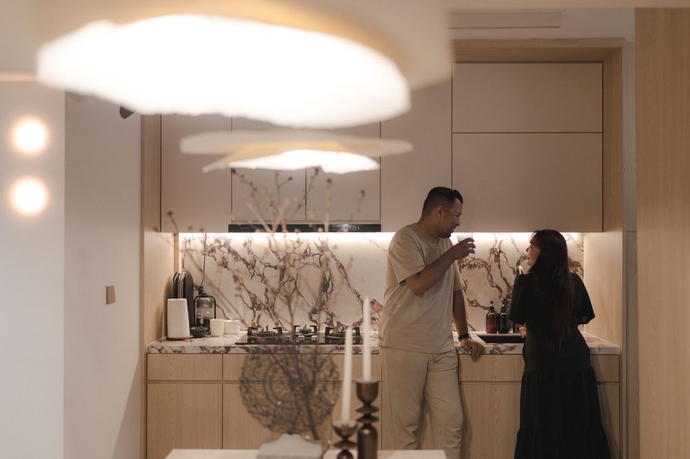 Two people stand in a modern kitchen with elegant pendant lighting and minimalist decor in warm neutral tones.