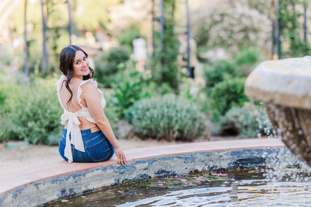 Someone sits at the edge of a garden fountain wearing a white top and blue jeans in a sunny outdoor setting.