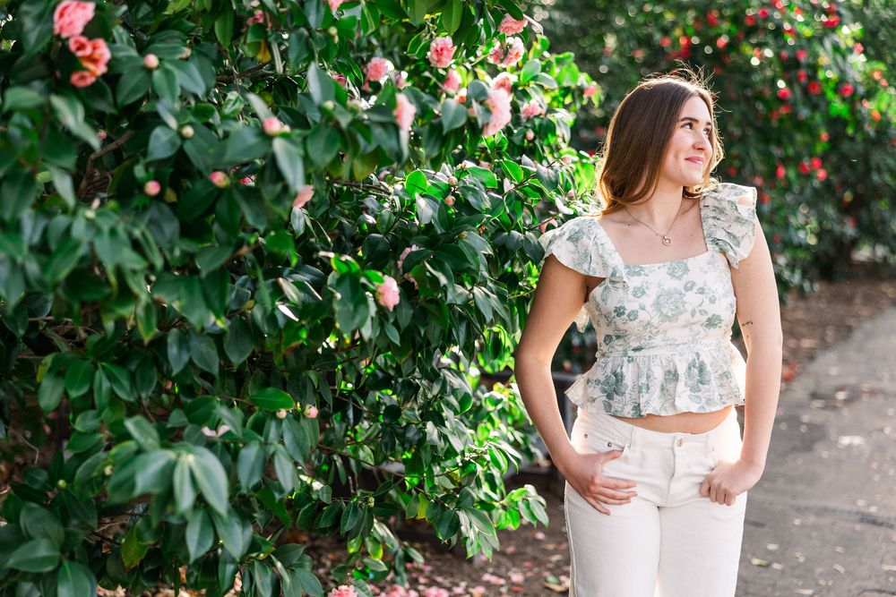 A woman in a floral crop top poses near pink blooming camellia bushes in a garden setting.