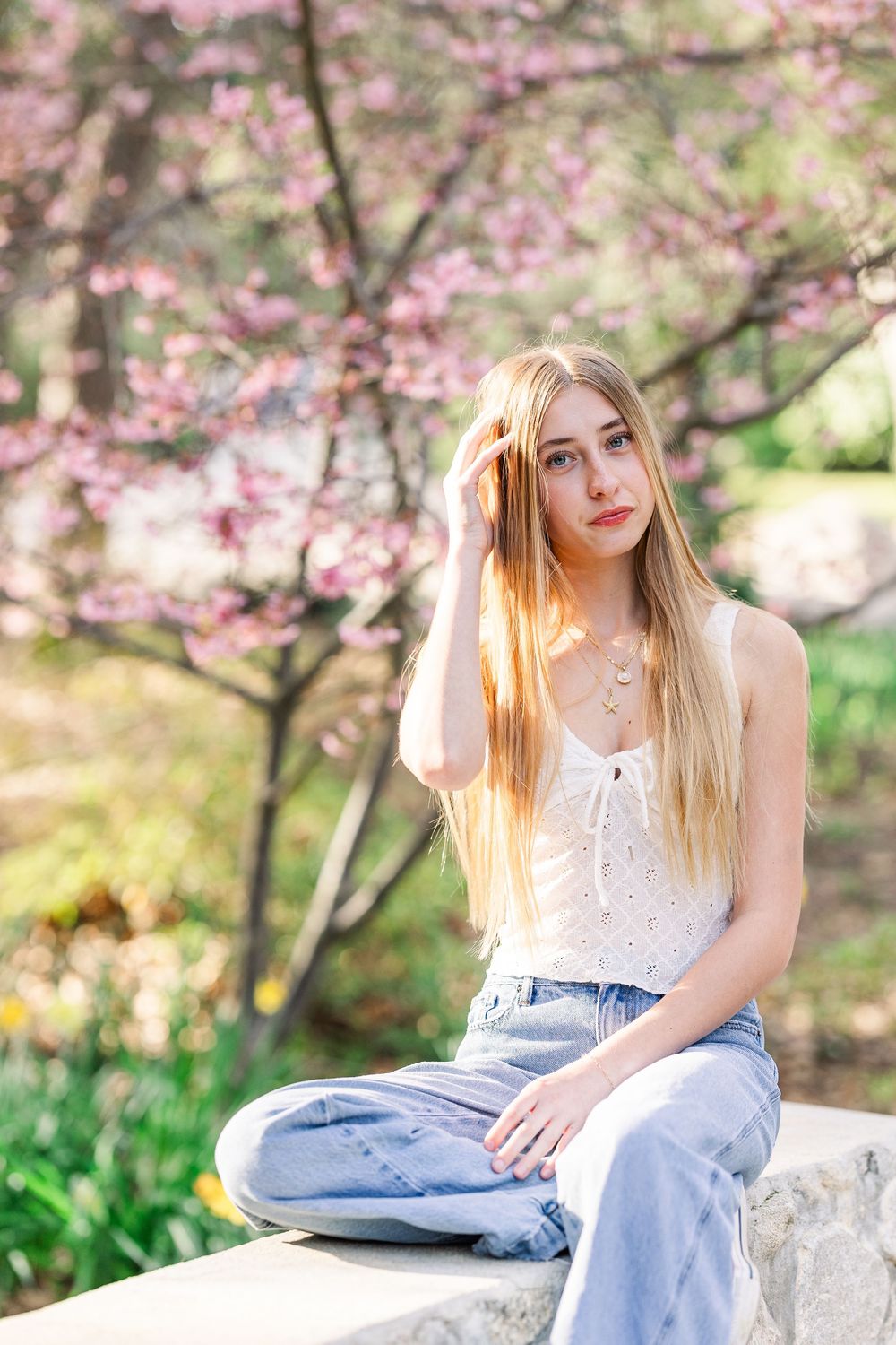 Casual spring portrait with cherry blossoms and white top with blue jeans seated on stone wall.