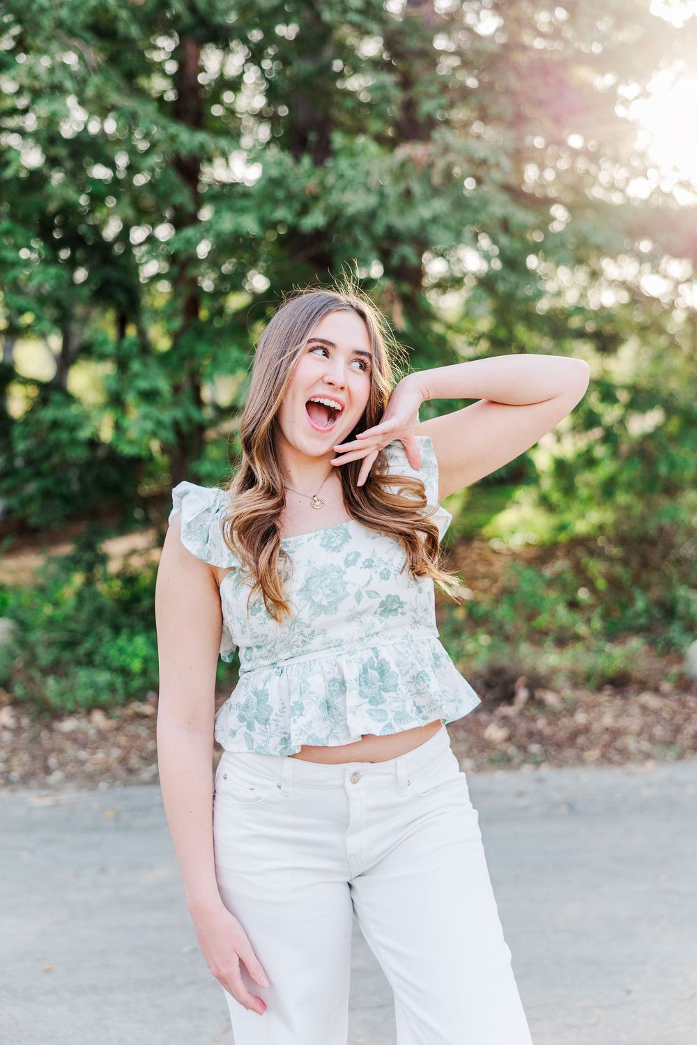 A joyful expression captured in casual summer attire with a floral peplum top and white pants against a sunlit green background.