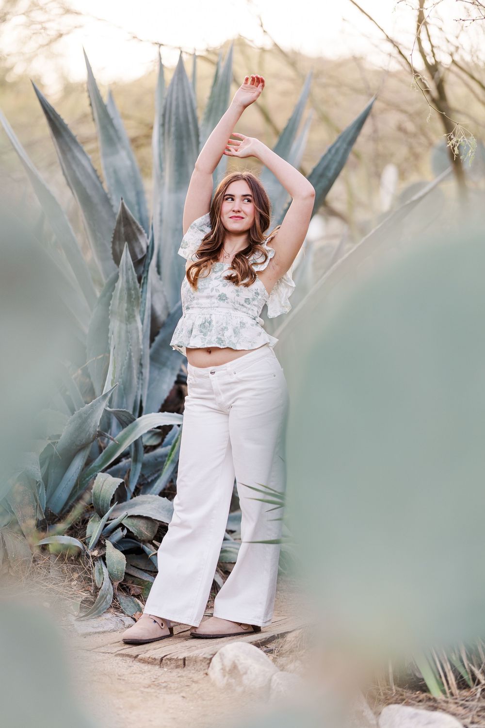 A woman in white attire poses gracefully among large agave plants in a desert garden setting at sunset.