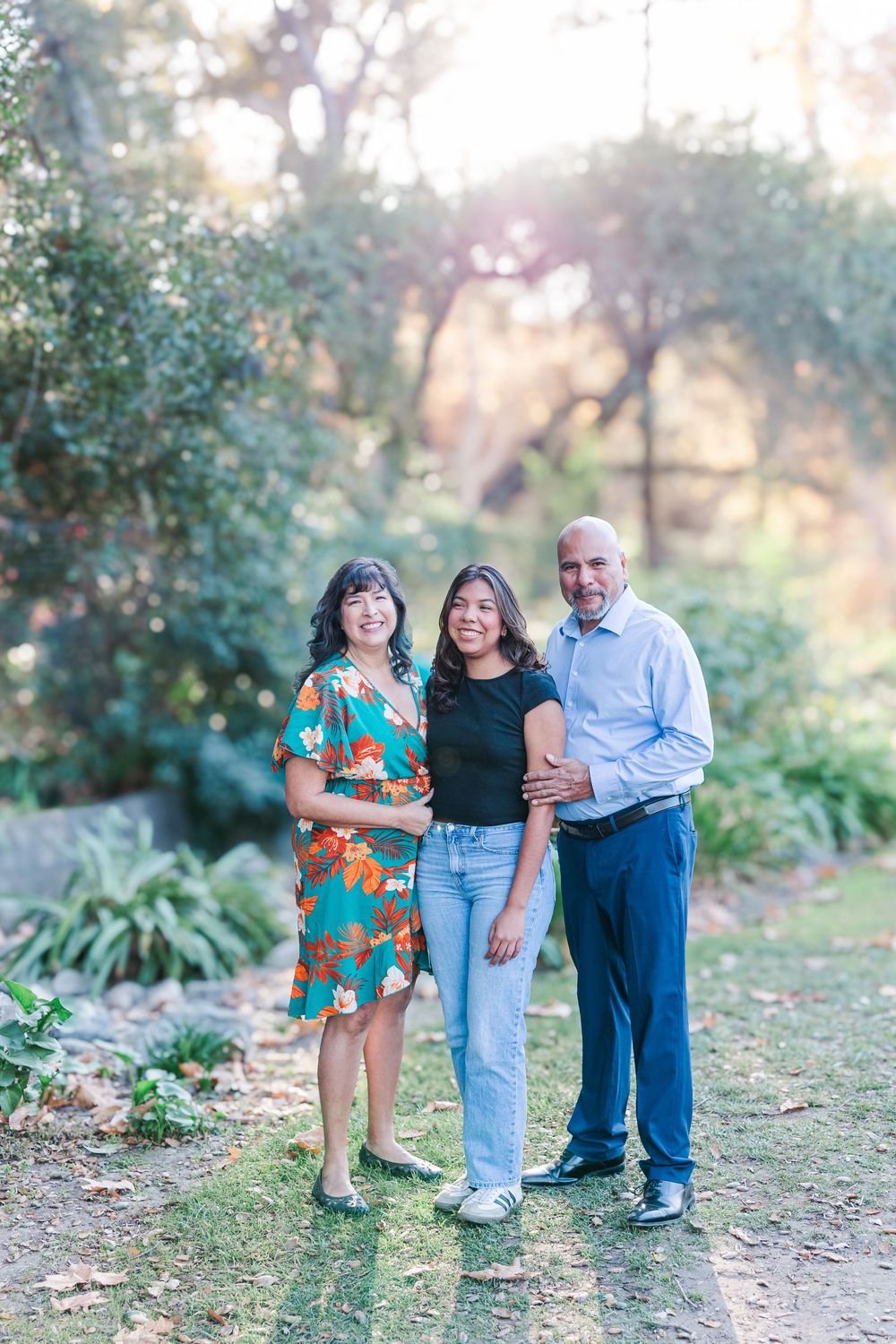 Three people stand together on a dirt path in a garden setting with trees and sunlight filtering through the background.