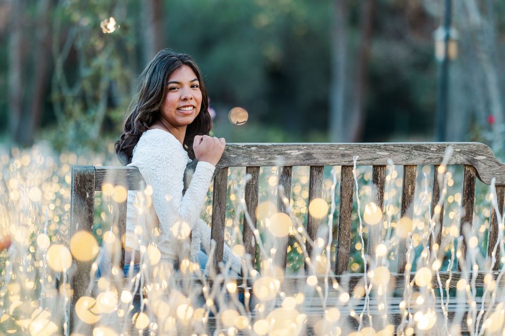 A young woman in a white sweater leans against a wooden railing surrounded by dreamy bokeh lights at sunset.