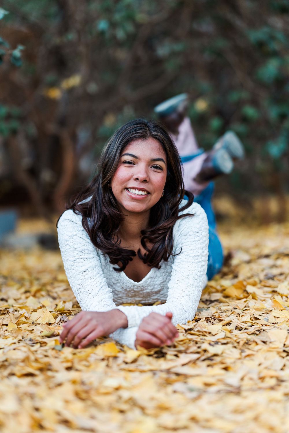 Person in white sweater lying on autumn leaves smiling while relaxing outdoors in natural lighting.