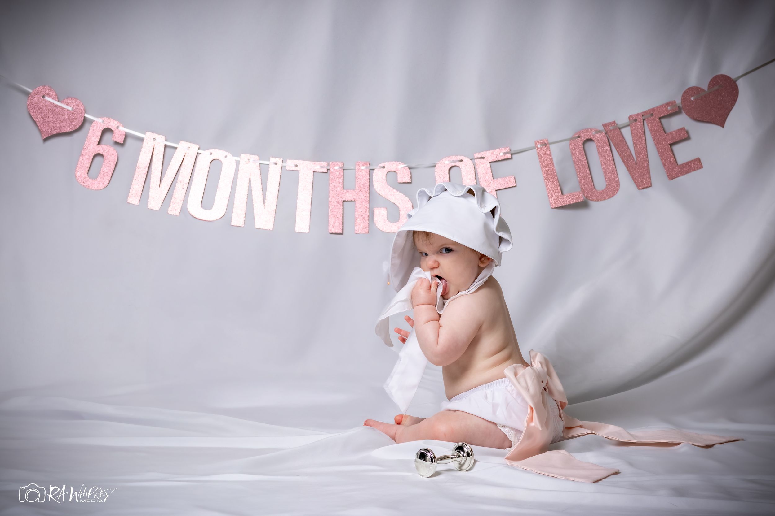 Baby in white outfit and crown sits under pink lettered banner against gray background.