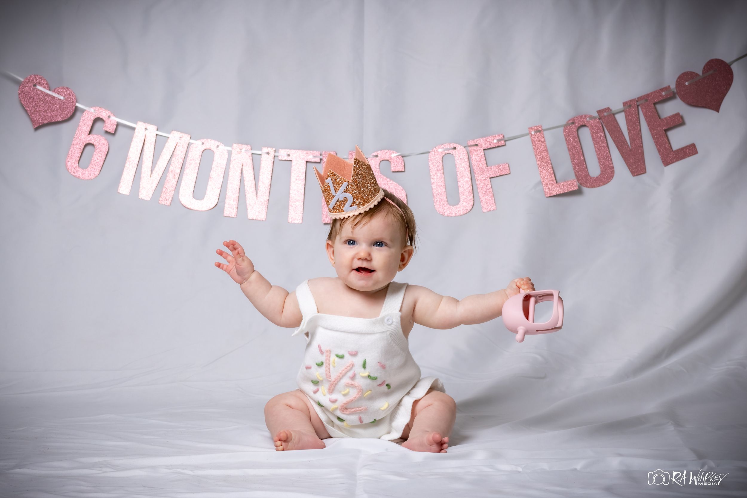 Baby wearing crown sits with arms outstretched under pink '6 months of love' banner on gray backdrop.