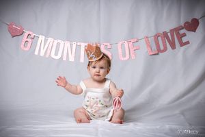 Baby in white outfit raises arm while sitting under decorative pink banner.