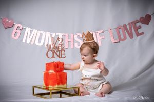 Baby reaches toward red tiered cake displayed on gold stand under pink banner.