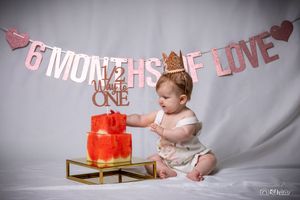 Baby explores red celebration cake on gold stand beneath pink banner.