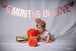 Baby in crown examines red tiered cake on gold stand under pink banner.