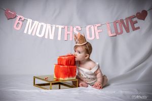 Baby sits beside red tiered cake under pink celebratory banner.